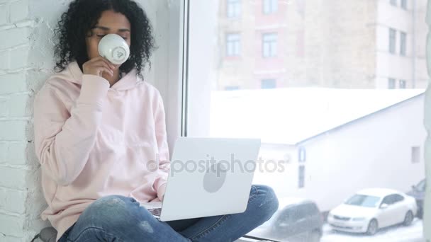 Mujer afroamericana bebiendo café y trabajando en computadora portátil, sentada en la ventana — Vídeos de Stock