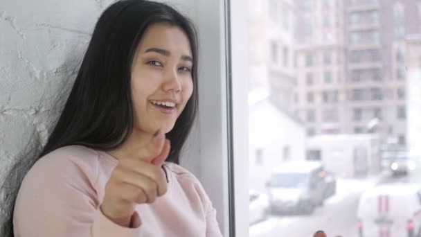 Joven mujer asiática señalando a la cámara, sentada en la ventana — Vídeos de Stock