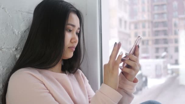 Mujer asiática joven navegando Smartphone, sentado en la ventana — Vídeo de stock