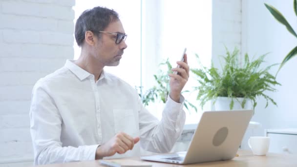 Chat de vídeo en línea por el hombre de mediana edad en el trabajo, Hablando en el teléfono inteligente — Vídeos de Stock