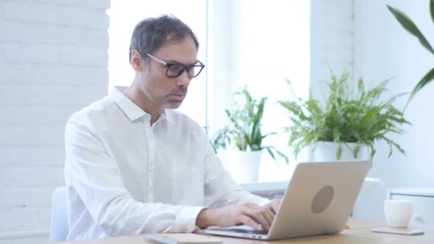 Mann mittleren Alters blickt während der Arbeit am Laptop im Büro in die Kamera — Stockvideo