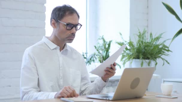 Чоловік середнього віку читає документи, сидячи в офісі — стокове відео
