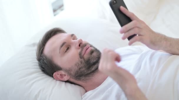 Веселый бородатый молодой человек дизайнер разговаривает по смартфону в постели — стоковое видео
