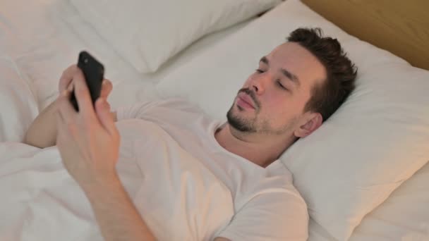 Молодой человек получает удар на смартфон в постели — стоковое видео