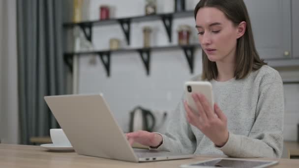 Akıllı telefon kullanan ve evde dizüstü bilgisayarda çalışan genç bir kadın. — Stok video