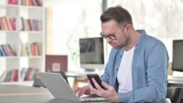 Mladý muž v brýlích pomocí chytrého telefonu a notebooku — Stock video