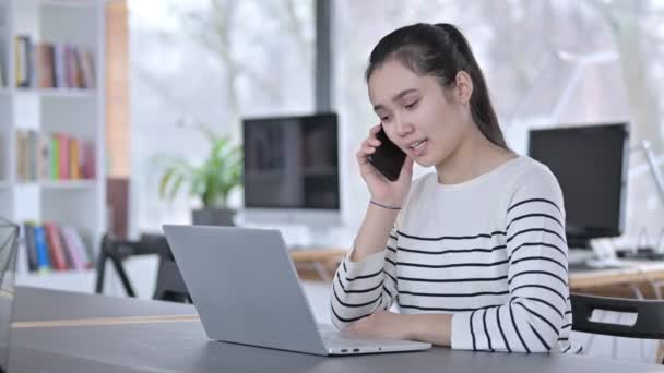 Junge Asiatin mit Laptop spricht im Büro mit Smartphone — Stockvideo