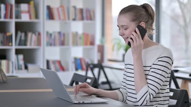 Jonge vrouw met laptop in gesprek op smartphone in de bibliotheek — Stockvideo