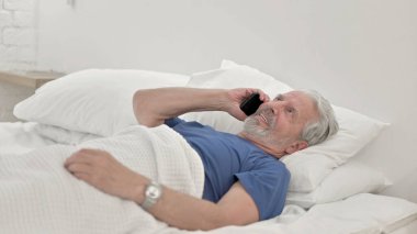 Yaşlı adam telefonda konuşuyor, yatakta yatıyor.