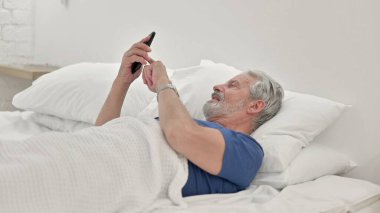 Yaşlı adam yatakta yatarken akıllı telefon kullanıyor.