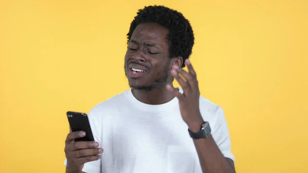 Casual African Man Reagera på förlust och användning av smartphone Isolerad på gul bakgrund — Stockfoto