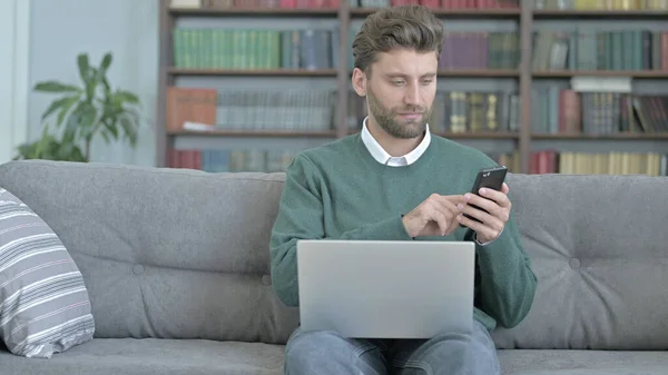 Трудолюбивый человек работает над ноутбуком, проверяя свой телефон — стоковое фото
