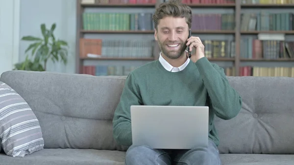 Молодой человек улыбается, разговаривая по телефону и работая над ноутбуком — стоковое фото