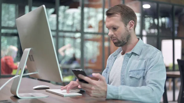 Kreativer junger Mann nutzt Smartphone und Computer bei der Arbeit — Stockfoto