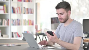 Genç adam ofiste akıllı telefon ve dizüstü bilgisayar kullanıyor.