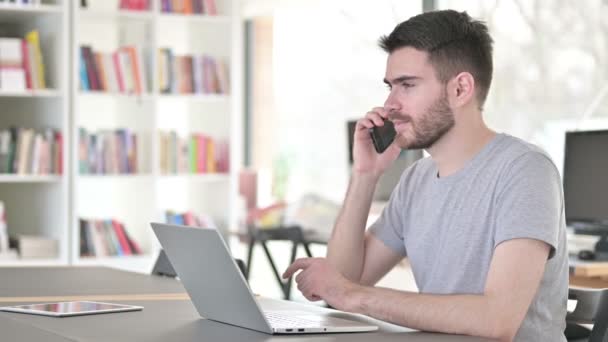 Jovem com Laptop Falando em Smartphone no Escritório — Vídeo de Stock