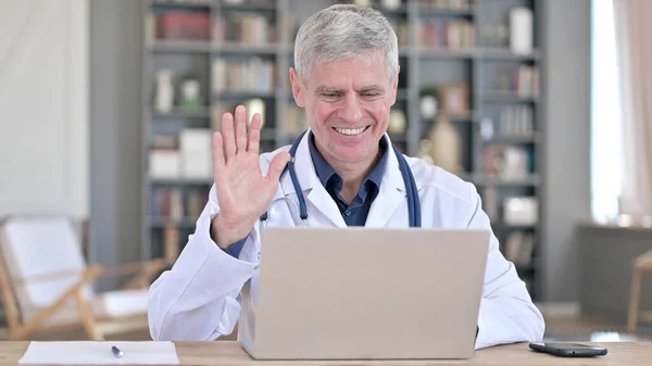Веселый старший врач делает видео-чат на ноутбуке, сидя в офисе — стоковое фото