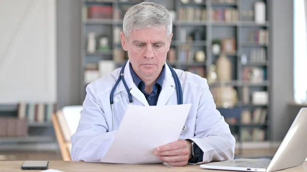 Médico de meia-idade profissional leitura de documentos enquanto sentado no escritório — Fotografia de Stock