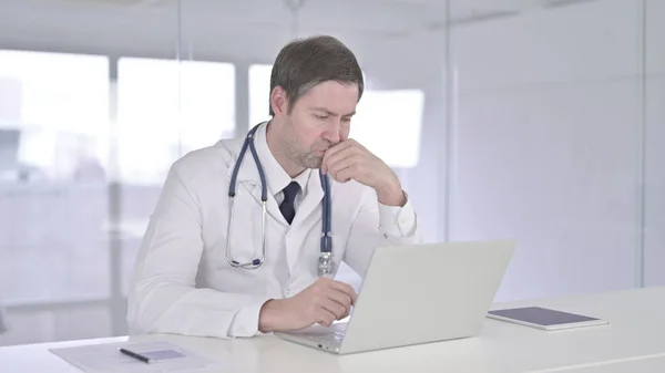 Medelålders läkare tänker och arbetar på laptop — Stockfoto