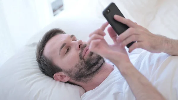 Hombre joven barba atractiva usando Smartphone en la cama —  Fotos de Stock
