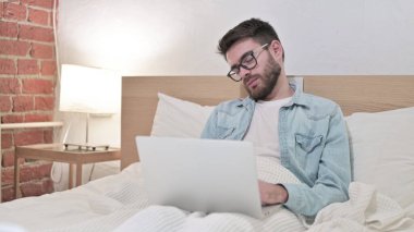 Gözlüklü genç adam dizüstü bilgisayarda çalışıyor ve yatakta kestiriyor.
