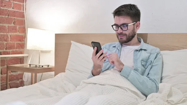 Привлекательный молодой человек в очках с помощью смартфона в постели — стоковое фото