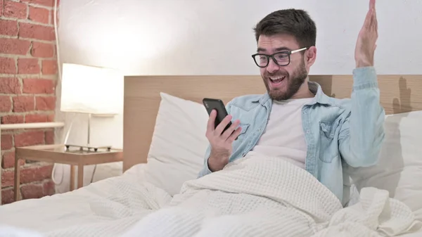 戴眼镜的年轻人在床上用智能手机庆祝成功 — 图库照片