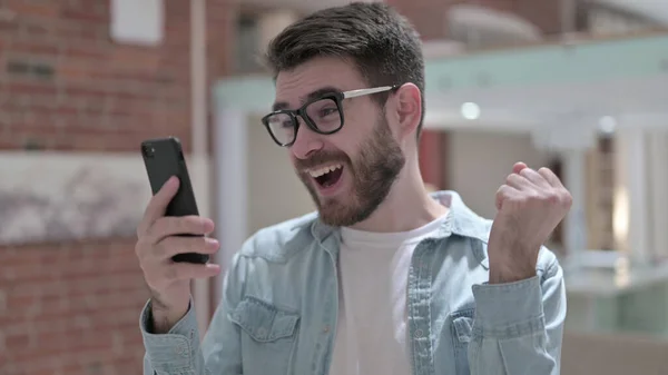 Vrolijke jonge man in glazen te vieren op de smartphone — Stockfoto