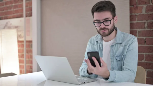 Mladý muž v brýlích pomocí chytrého telefonu a práce na notebooku — Stock fotografie