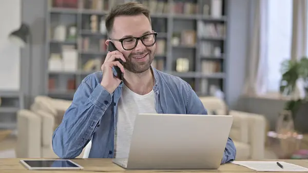 Junger Mann spricht auf Smartphone und arbeitet am Laptop — Stockfoto