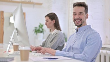 Genç Yaratıcı Adam Masaüstünde çalışıyor ve Kameraya Gülümsüyor