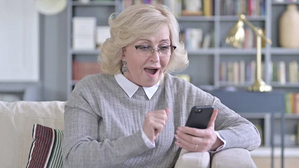 Oude vrouw viert succes op smartphone op de bank — Stockfoto
