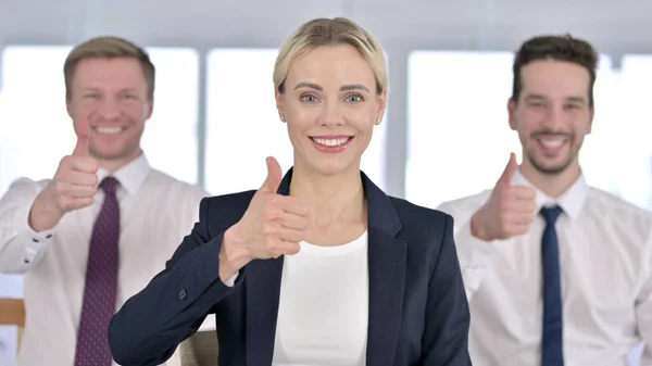 Portrét atraktivní podnikatelky stojící s týmem a ukazující palce nahoru — Stock fotografie