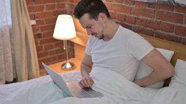 Yatakta dizüstü bilgisayarla uğraşırken sırt ağrısı çeken genç adam.