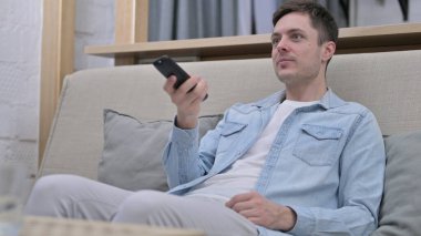 Genç adam TV izlerken koltukta oturuyor, kanal değiştiriyor.