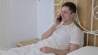 Genç adam yatakta otururken telefonda konuşuyor.