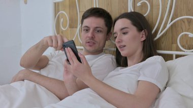Yatakta akıllı telefon kullanan neşeli genç çift.