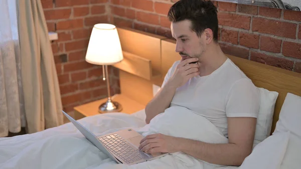 Jongeman denken en werken op laptop in bed — Stockfoto
