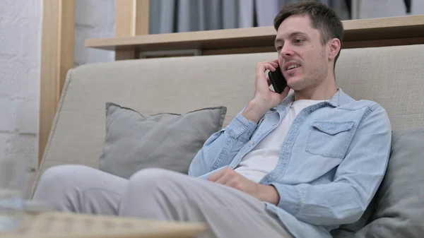 Молодой человек сидит на диване и разговаривает по смартфону — стоковое фото