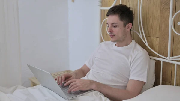 Hardwerkende jongeman werkt op laptop in bed — Stockfoto