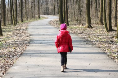 Kırmızı ceketli ve kırmızı şapkalı küçük kız asfalt yoldaki ağaçların arasında bahar ormanlarında yürüyor.