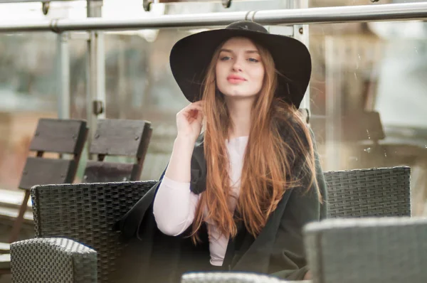 Girl in a black wide-brimmed hat and cloak sitting on the terrace of the restaurant — Stock Photo, Image