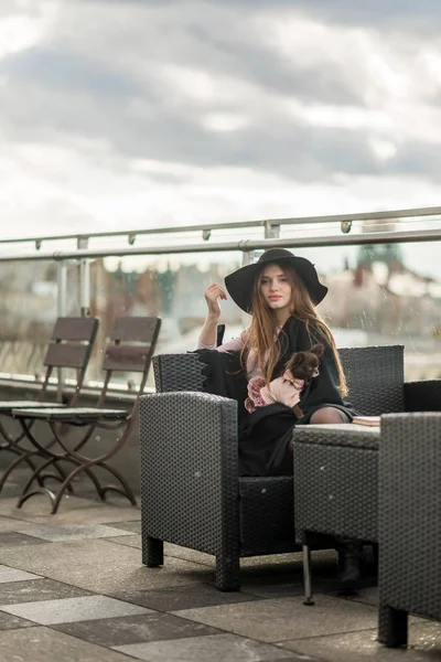 Chica en un sombrero de ala ancha negro y capa sentado en la terraza del restaurante — Foto de Stock