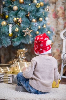 Kırmızı şapkalı bir çocuk bir Noel ağacı bir hediye ile oturuyor.