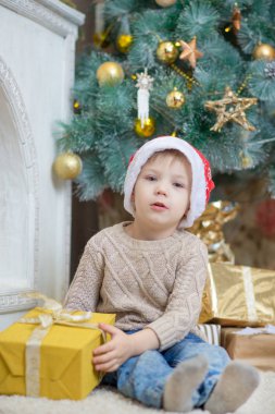 Kırmızı şapkalı bir çocuk bir Noel ağacı bir hediye ile oturuyor.
