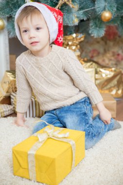 Kırmızı şapkalı bir çocuk bir Noel ağacı bir hediye ile oturuyor.