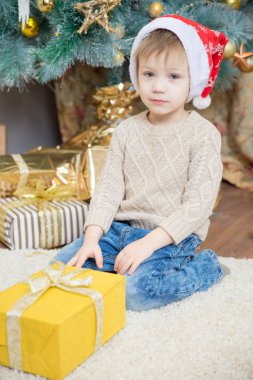 Kırmızı şapkalı bir çocuk bir Noel ağacı bir hediye ile oturuyor.