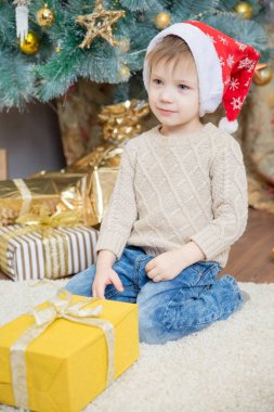 Kırmızı şapkalı bir çocuk bir Noel ağacı bir hediye ile oturuyor.