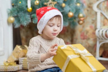 Kırmızı şapkalı bir çocuk bir Noel ağacı bir hediye ile oturuyor.