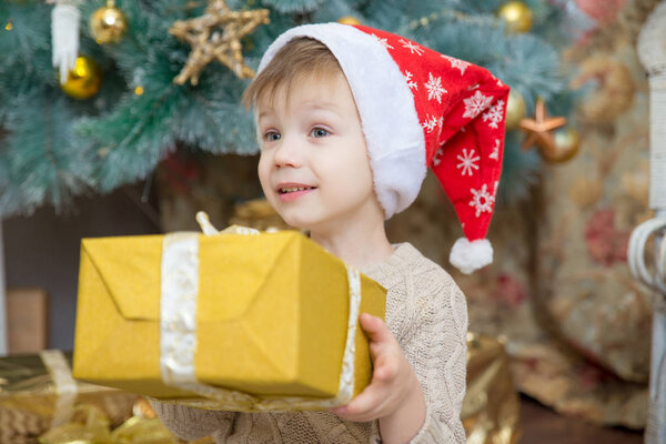 a boy in a red hat sits at a Christmas tree with a gift